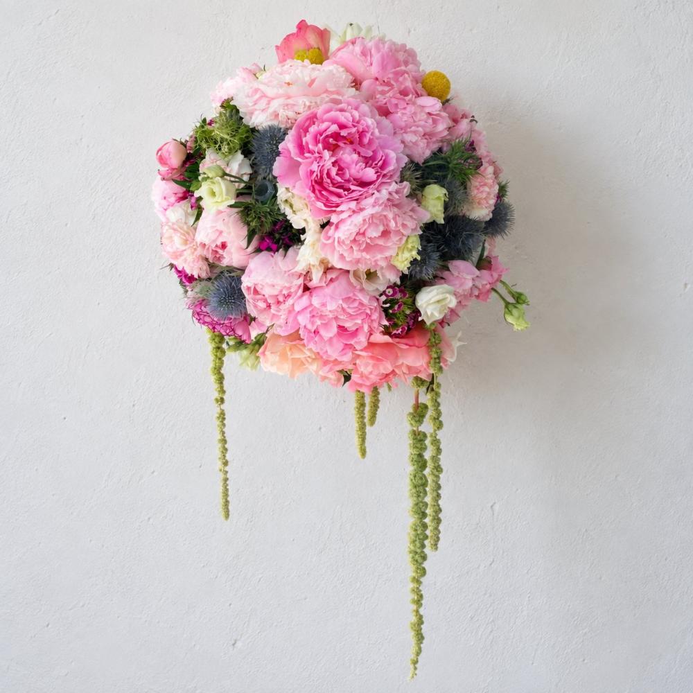 Hanging floral arrangement with pink peonies, ranunculus, and trailing green amaranthus against a white wall