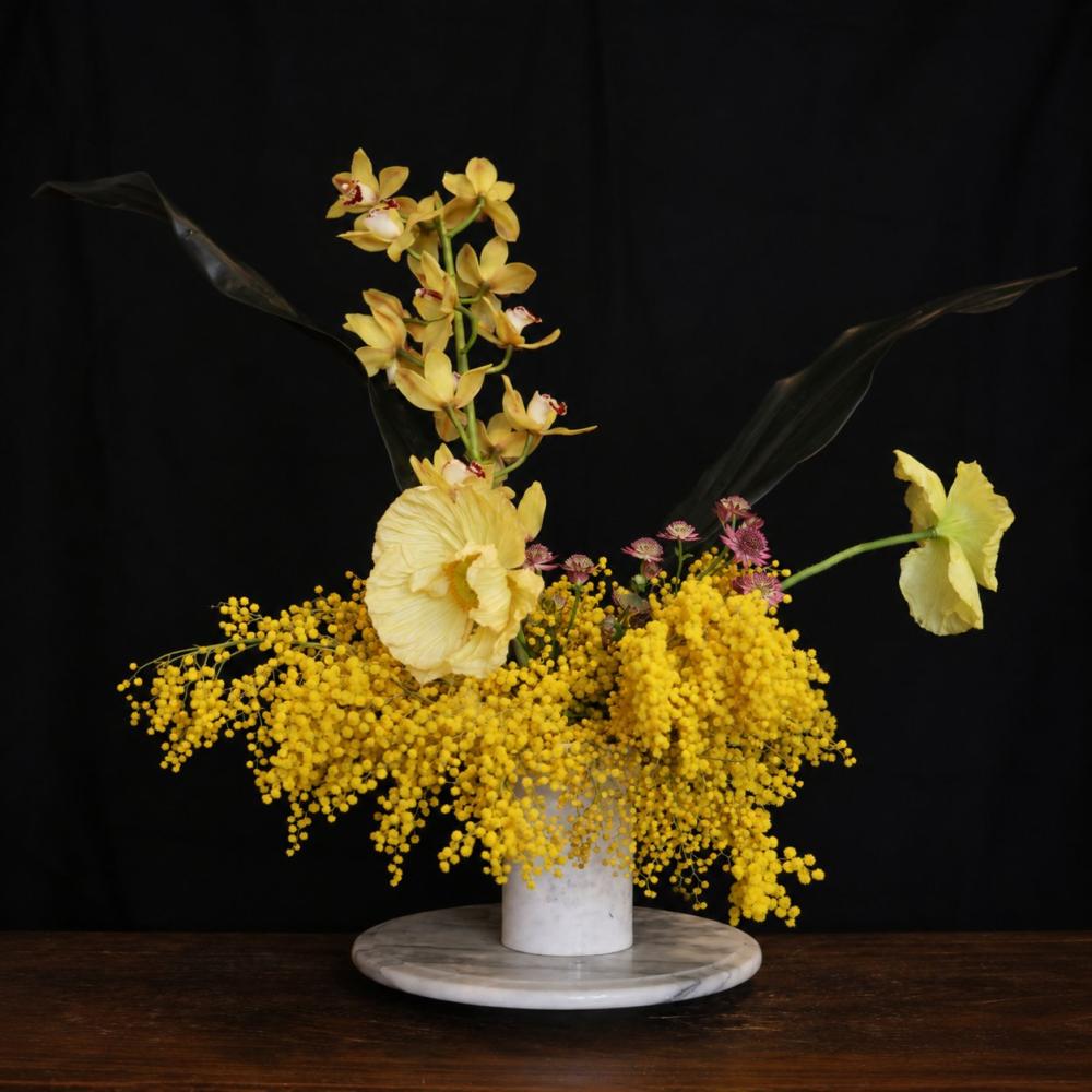 Yellow mimosa, cymbidium orchids, and pale poppies on a white marble stand against a black backdrop at Royal Highness, Fitzroy, Berlin