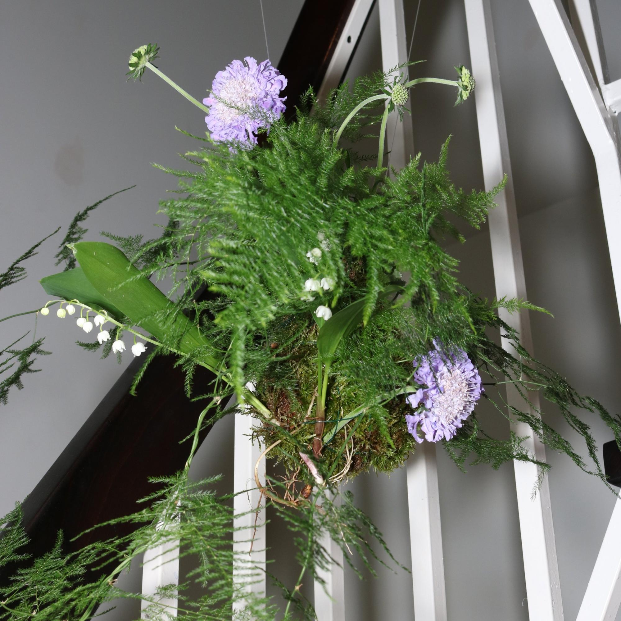 Hanging floral arrangement with purple scabiosa, ferns, and lily of the valley suspended against a white stair railing